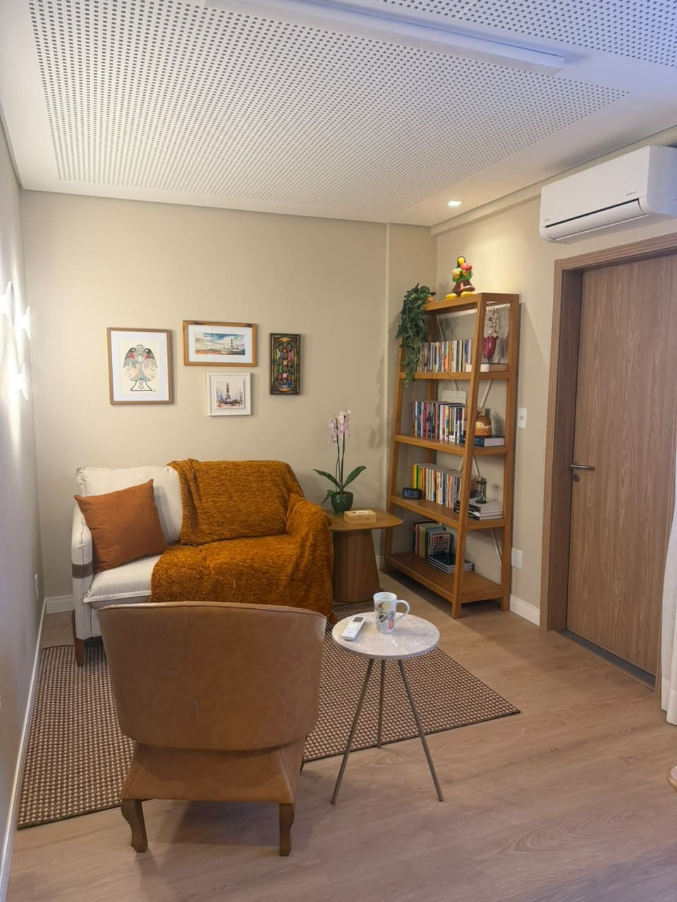 Sala de atendimento do consultório em Aracaju, com poltronas, manta caramelo, estante de livros e quadros na parede